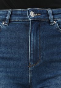 Blå denimjeans med hög midja, med en silverknapp, synliga sömmar och en slät, strukturerad yta.