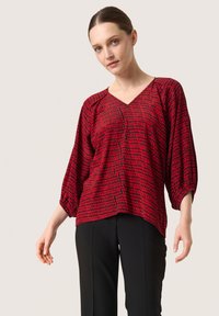 Rød tunika top med V-hals, batwing ærmer og et tekstureret kvadratmønster. Stoffet ser let ud og falder løst.