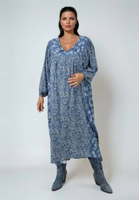 Lange blauwe jurk met een paisleypatroon, V-hals en geplooide buste. De stof is zacht, met lange mouwen, gecombineerd met grijze enkellaarsjes.