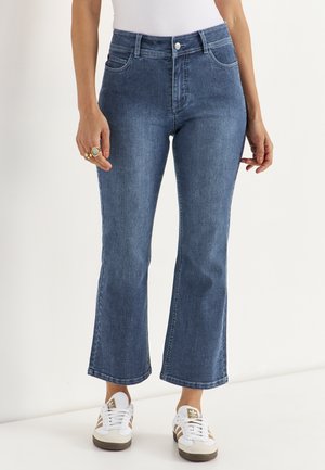Kvinde iført blå denim cropped flare jeans med middel talje og hvide sneakers med beige striber, stående mod en hvid baggrund.