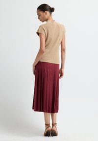 Top de punto acanalado beige con mangas cortas, combinado con una falda midi plisada burdeos. Mostrado desde la parte de atrás, con sandalias de tacón marrones.