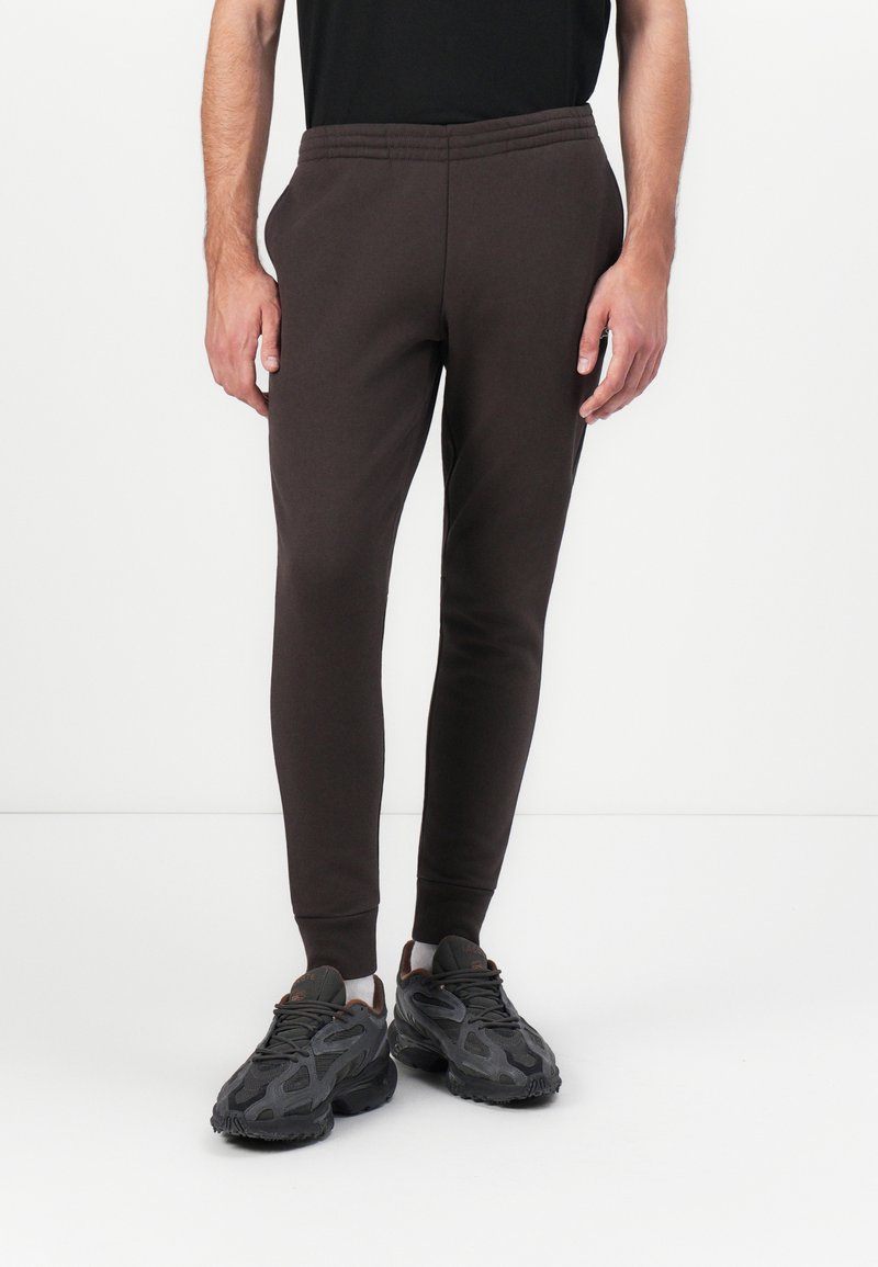 Braune, schmal zulaufende Jogginghose aus weichem Stoff mit elastischem Bund und Bündchen, kombiniert mit klobigen schwarzen Sportschuhen.