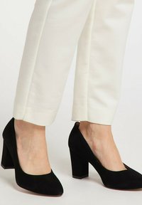 Füße, die schwarze Wildleder-Blockabsatzpumps tragen, kombiniert mit cremefarbenen, cropped Hosen vor einem schlichten Hintergrund.