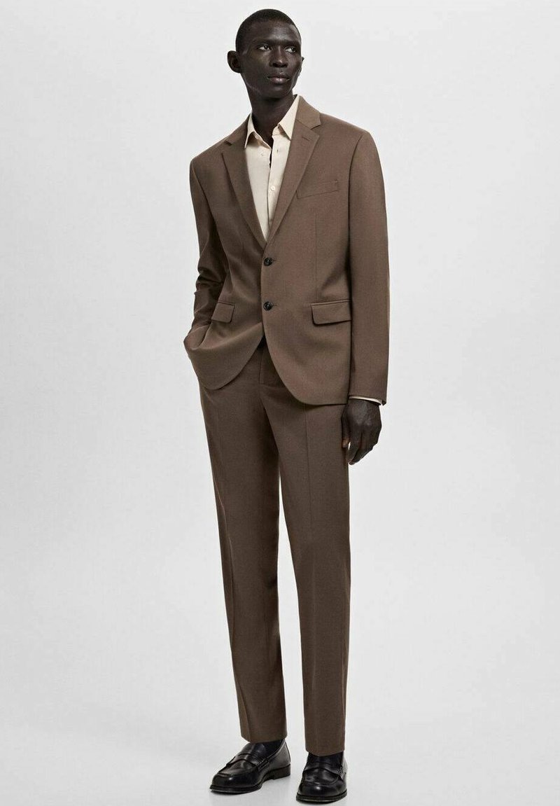 Costume marron sur mesure en tissu lisse avec une veste à simple boutonnage, des revers à crans et un pantalon à plis plats, associé à une chemise claire.