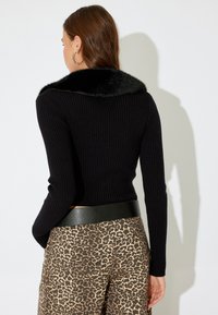 Schwarzer gerippter Pullover mit einem Kragen aus Kunstfell, kombiniert mit braunen Hose im Leopardenmuster und einem breiten schwarzen Gürtel. Der Stoff wirkt weich und strukturiert.