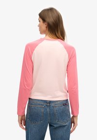 Vrouw met lang haar die een lichtroze en donkerroze raglan longsleeve en een blauwe spijkerbroek draagt, met haar rug naar de camera toe.