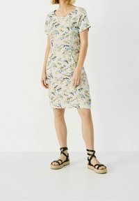 Vestido de estampado floral en color beige claro con mangas cortas, que presenta detalles en azul y amarillo. Llevado con sandalias negras de tiras y suela de esparto natural.