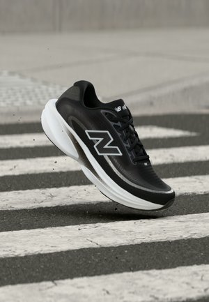 Fekete-fehér New Balance futócipő a levegőben, egy gyalogátkelő felett, aszfaltúton.