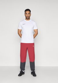 Λευκό αθλητικό μπλουζάκι με μαύρο λογότυπο Nike, συνδυασμένο με κόκκινες και γκρίζες παντελόνες που φέρουν λογότυπο. Μαύρα παπούτσια ολοκληρώνουν την εμφάνιση.