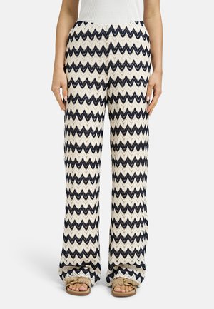 Woman wearing wide-leg pants with black and white zigzag pattern and tan sandals, standing against white background.