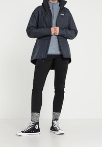 Wodoodporna ciemnoszara kurtka z kapturem na jasnoszarym topie na suwak, połączona z czarnymi spodniami i czarnymi sneakersami za kostkę.