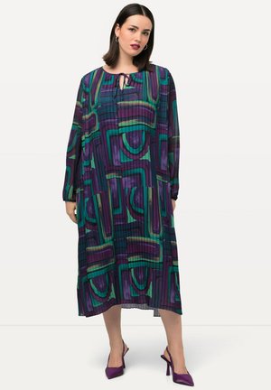 Langärmliges, plissiertes Kleid mit einem geometrischen Muster in Lila, Grün und Blau, Schlüssel-Ausschnitt und lockerer Silhouette. Kombiniert mit lila High Heels.