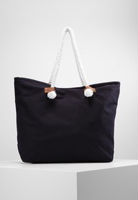 Marinblå canvas tote bag med vita rephandtag, med bruna läderdetaljer och pom-poms. Stort, ostrukturerat design för gott om förvaring.