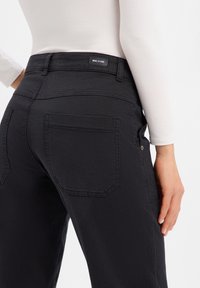 Jean en denim noir à coupe ajustée, avec deux poches arrière et une étiquette sur la ceinture. Le tissu présente une légère texture.