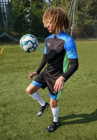 Schwarzes Fußballtrikot mit kurzen Ärmeln und blauen sowie grünen Akzenten, mit einem Reißverschluss am Hals. Schwarze Fußballschuhe und weiße Socken auf einem Kunstrasenplatz.
