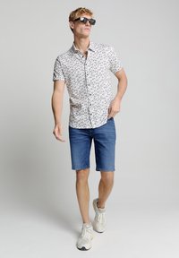 Korte mouw overhemd met een bladerenpatroon, witte achtergrond; gecombineerd met denim shorts en lichte sneakers. Zwarte zonnebril op.