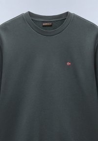 Sudadera gris oscuro de algodón. Presenta un cuello acanalado y un pequeño detalle de bandera bordado en el pecho. Textura suave.