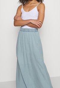 Kvinna med korslagda armar som bär en ärmlös vit topp och en högmidjad ljusblå maxikjol mot en enkel bakgrund.