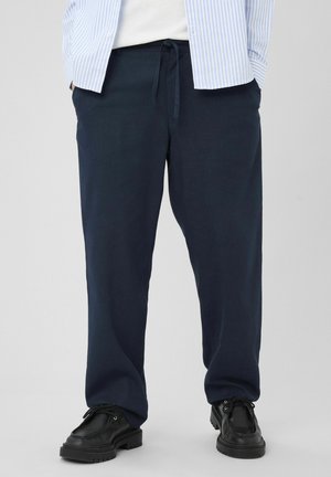 Homme portant un pantalon à cordon de serrage bleu marine, des chaussures en cuir noires, une chemise blanche et une surchemise rayée bleu et blanc, debout sur un fond gris clair.