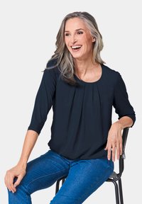 Marineblaue Bluse mit Dreiviertelärmeln, runder Halslinie und Faltendetail an der Vorderseite, kombiniert mit blauen Jeans.