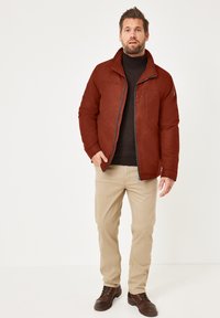 Rote gesteppte Jacke mit Stehkragen, Fronttaschen und Reißverschluss, kombiniert mit hellbeigen Hosen und braunen Schnürstiefeln.