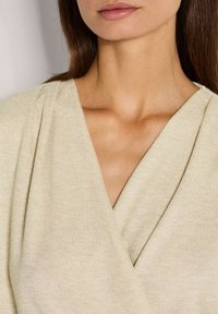 Beige Wickeloberteil aus strukturiertem Strickstoff mit tiefem V-Ausschnitt und drapiertem Design, das einen subtilen Schimmer hervorhebt.