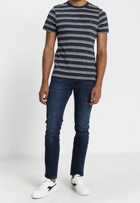 Maglietta a righe navy con linee orizzontali bianche, collo a giro, abbinata a jeans slim fit blu scuro e scarpe da ginnastica bianche con dettagli neri.