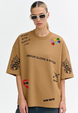 Femme portant un t-shirt oversize beige avec texte noir, patchs emoji colorés, illustrations de dés et lunettes de soleil blanches rectangulaires.