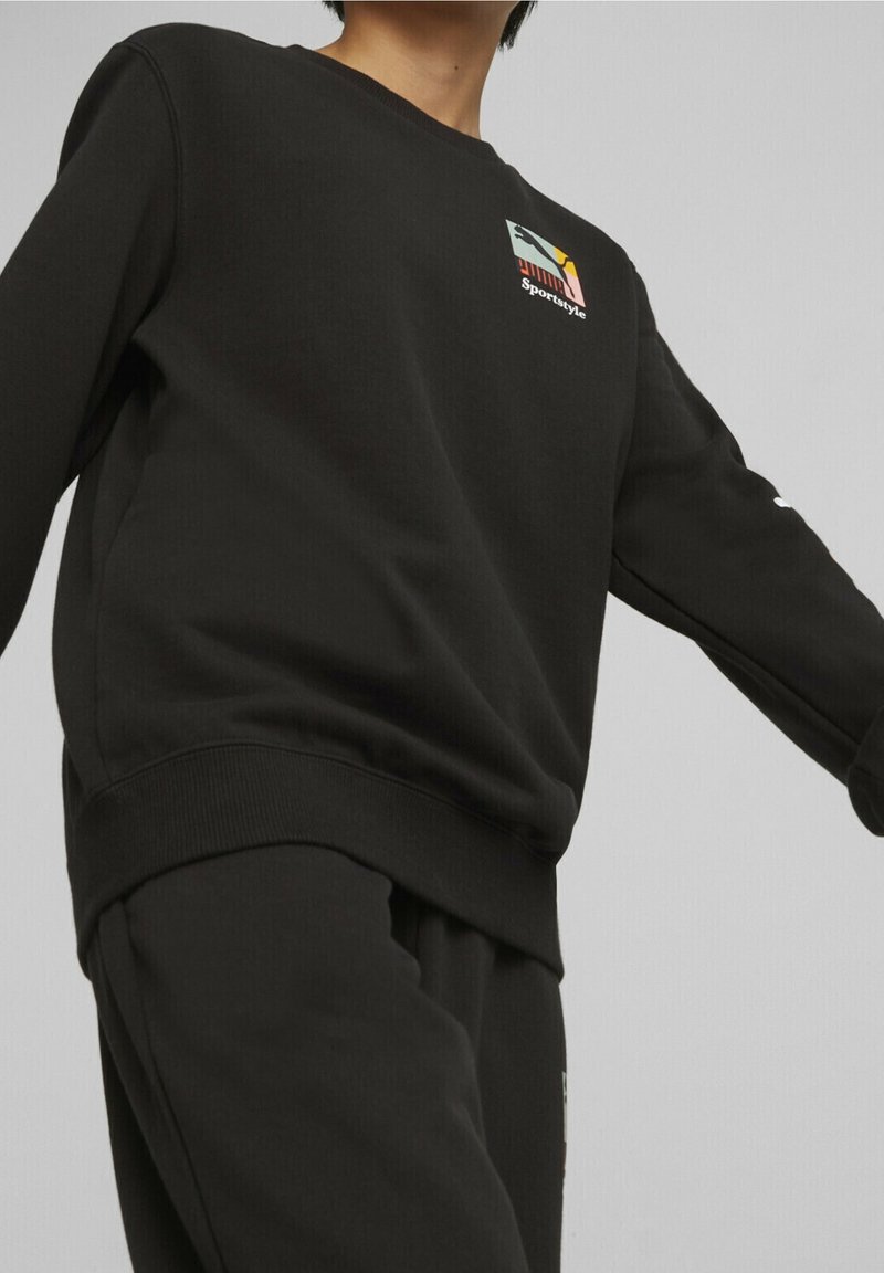 Personne portant un sweat-shirt Puma Sportstyle noir et un pantalon noir assorti sur un fond gris uni.