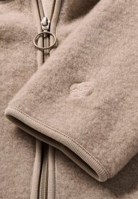 Giacca in pile beige con zip, caratterizzata da polsini a coste, un cursore della zip rotondo e un delicato dettaglio di ricamo floreale sulla manica.