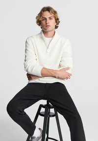 Pullover bianco a maglia con colletto a mezza zip, dotato di polsini e orlo a coste. Indossato con jeans scuri e sneakers grigie mentre è seduto su uno sgabello.