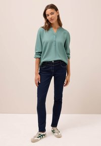 Grön V-ringad knapp-up blus i kombination med mörkblå skinny jeans och grå-gröna sneakers med gröna accenter. Slät tygkvalitet och åtsittande design.