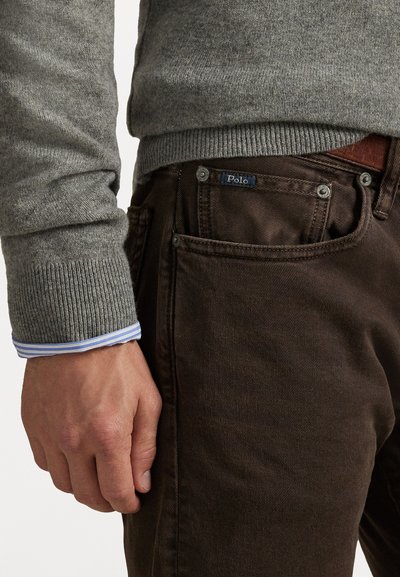 Pullover griș cu dungi, purtat peste pantaloni maro, având eticheta Polo vizibilă. Manșeta unei cămăși în dungi albastre și albe iese în evidență de sub pulover.