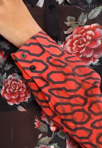 Blusa rossa a fantasia con maniche che presenta un motivo esagonale nero con dettagli a bottoni, abbinata a uno sfondo in tessuto trasparente floreale nero.