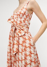 Cvjetna haljina s bijelom pozadinom i uzorcima crvenog i zelenog lišća, prilagođeni korpus, volanasti dekolte, vezanje u struku i bočne džepove.