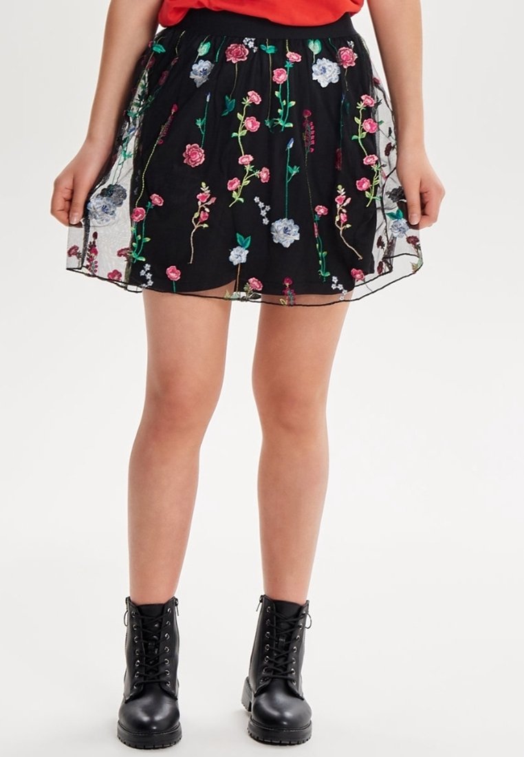 Jupe noire avec broderie florale en rose, bleu et vert. Superposition transparente et taille plissée. Associée à des bottes noires à lacets.