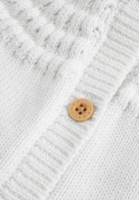 Bela pletena cardigan z teksturiranim rebrastim vzorcem in dekorativnim bambusovim gumbom z vgraviranim napisom "NEXT". Mehak, teksturiran detajl tkanine.