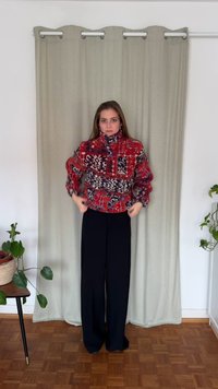 Czerwona kurtka z polaru z wzorami i wysokim kołnierzem, zestawiona z czarnymi spodniami o szerokich nogawkach. W tle jasna zasłona i rośliny doniczkowe.