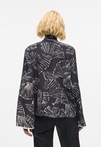 Fekete blúz fehér levélmintával, puha anyagból, hosszú ujjakkal és laza szabással. Alternatív nyílás magas nyakkivágással.