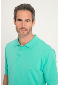 Turkos polo skjorta med en mjuk textur, treknappsstängning och en broderad logotyp på vänster bröst. Korta ärmar.