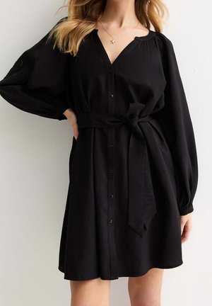 Kvinde iført en sort kjole med knapper, lange pufærmer og bælte bundet om taljen, stående mod en ensfarvet lys baggrund.