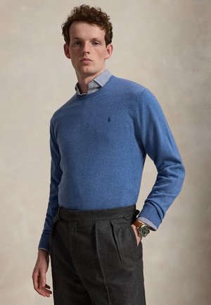 Homme aux cheveux bouclés portant un pull bleu sur une chemise rayée, un pantalon plissé foncé et une montre-bracelet avec un bracelet en cuir marron.