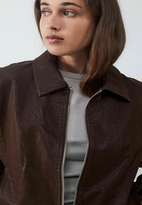 Brązowa, strukturalna skórzana kurtka z kołnierzykiem i srebrnym zamkiem, noszona na jasnoszarym koszuli. Skup się na designie i materiale kurtki.