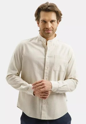 Mężczyzna z jasnobrązowymi włosami, ubrany w beżową koszulę zapinaną na guziki z kołnierzykiem typu mandarynka, trzymający ręce splecione z przodu na jednolitym tle.