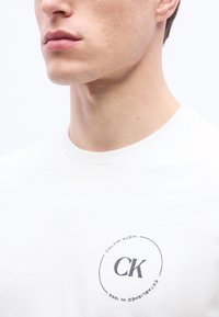 Άνδρας που φοράει λευκό T-shirt Calvin Klein με κυκλικό λογότυπο "CK" και "Established in 1968" στο στήθος, μπροστά σε απλό φόντο.