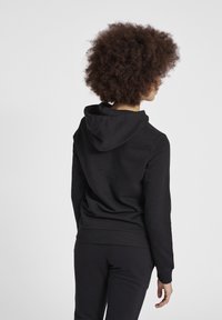 Felpa nera con cappuccio, dalla consistenza morbida, caratterizzata da una vestibilità comoda e polsini a costine, vista di schiena. Indossati dei pantaloni neri abbinati.