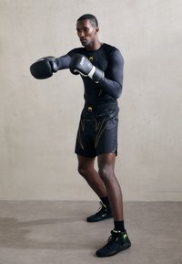 Gants de boxe noirs, t-shirt ajusté à manches longues noirs avec des accents jaunes, shorts noirs avec un design graphique, et chaussures noires avec des détails néon.