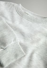 Světle šedá mikina s žebrovaným kulatým výstřihem a manžetami; vyrobena z jemné, strukturované tkaniny s jemnými barevnými variacemi.