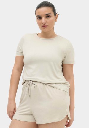 Kvinde med mørkt hår i en lav knold iført en beige t-shirt med korte ærmer og matchende shorts med snøre, standing mod en ensfarvet lys baggrund.