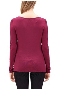 Kobieta w długim, różowym topie i czarnych spodniach, stojąca tyłem na białym tle.